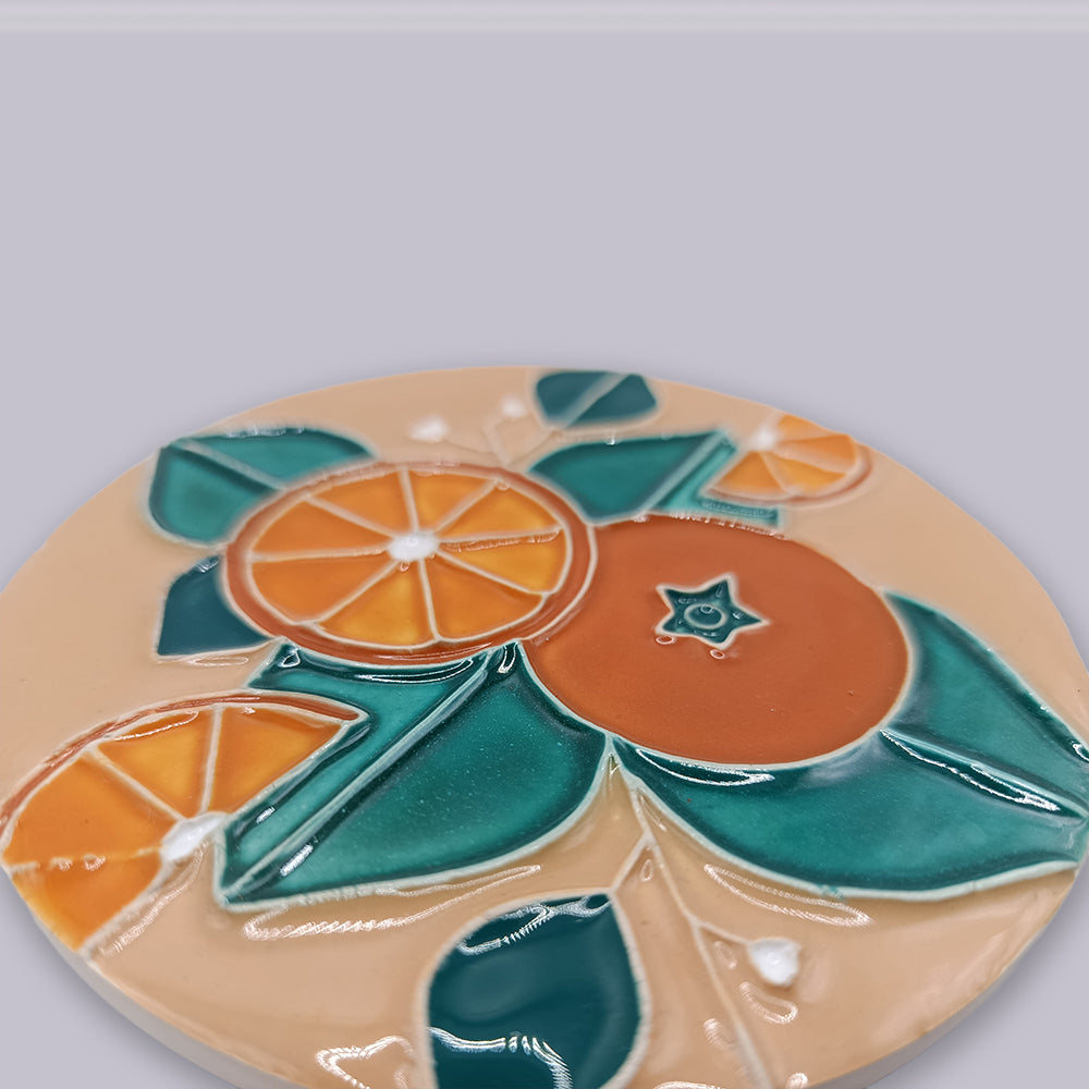 The Algarve Sandstone Orange Ceramic Coaster by Miradouro Ceramics features a glossy finish with vibrant orange slices, a whole orange, and green leaves set on a beige background.