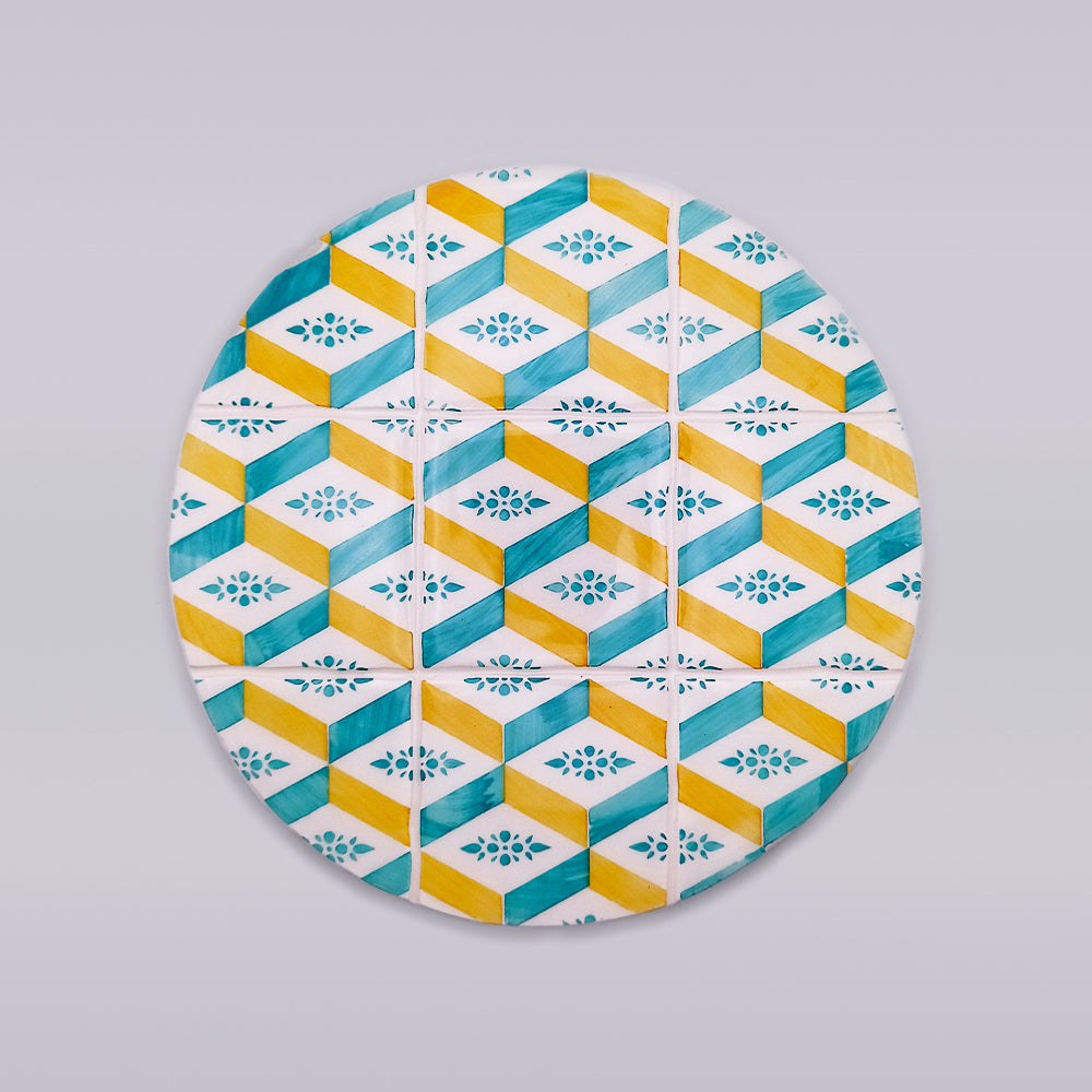 The Arraiolos Ceramic Tile Trivet by Tejo Shop showcases a geometric pattern with blue, yellow, and white cubes and floral motifs. This circular wall art design is divided into four vertical sections, creating a tiled effect against a plain light gray background.