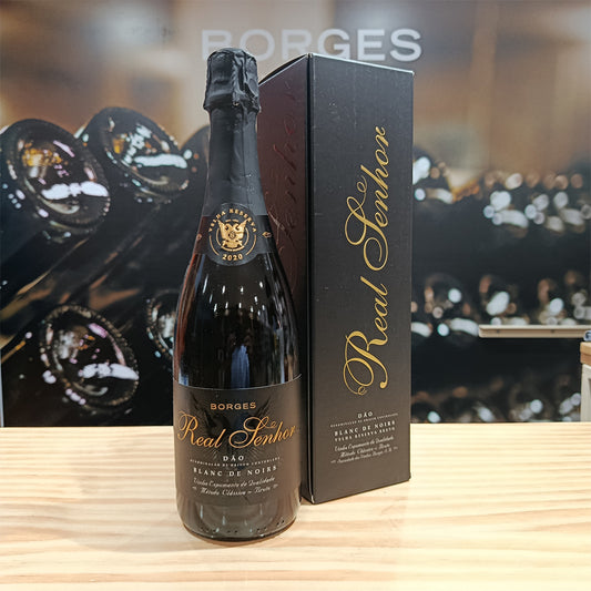 A bottle of Borges Wines Real Senhor Blanc de Noirs Old Reserv Brut stands next to its black and gold box on a wooden surface, with blurred bottles—including Borges Real Senhor Blanc de Blancs—visible in the background.