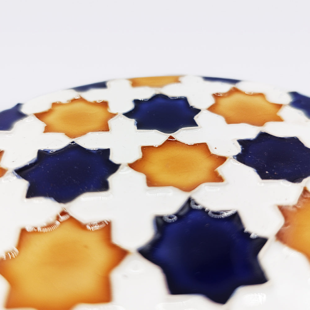 Close-up of the Heritage Star Mosaic Coaster by Miradouro Ceramics, featuring a glossy, hand-painted Mudéjar star pattern in navy blue and amber with white lines. The azulejo-inspired surface reflects light; the bright background is softly blurred.