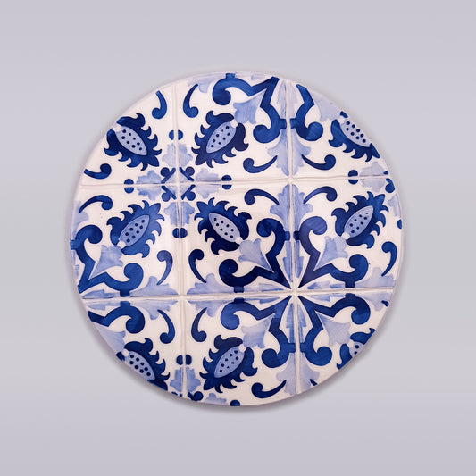 A hand-crafted Tejo Shop Sagres Ceramic Pad for Hot Pans showcases a circular decorative tile with a repeating pattern of blue floral and abstract designs on a white background, creating an intricate and symmetrical motif across the surface. This exquisite piece is meticulously hand-painted, adding a touch of artisanal elegance to any space.