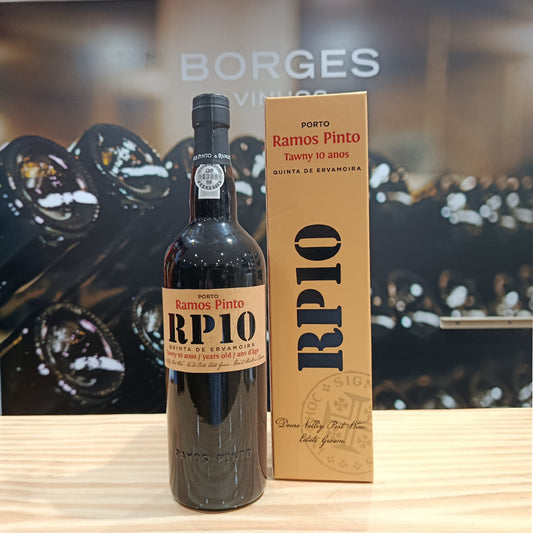 A bottle of Ramos Pinto 10 Year Old Tawny Porto - Quinta de Ervamoira stands beside its beige box on a wooden surface, made from Douro Demarcated Region grapes, with blurred wine bottles in the background.