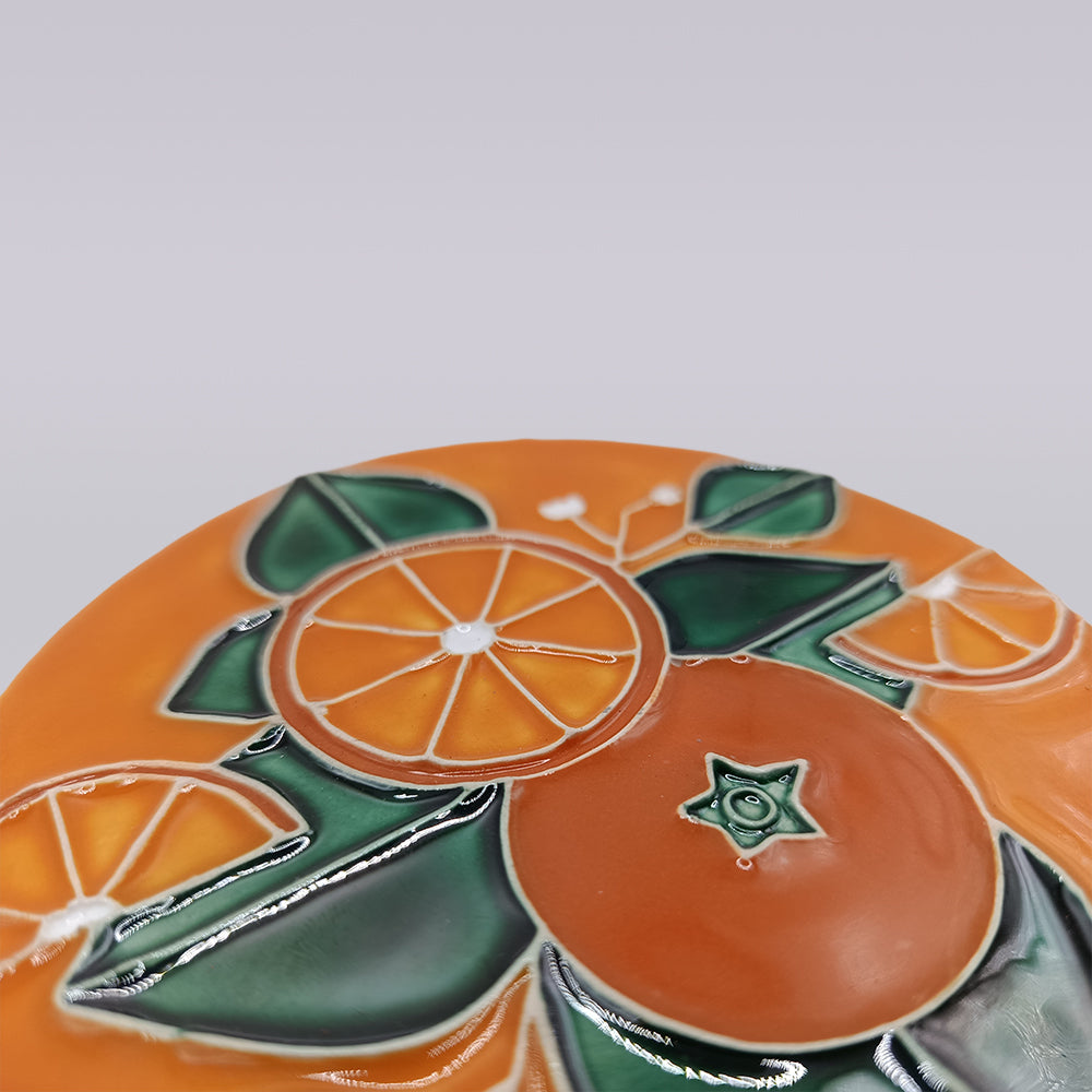 A close-up of the Algarve Orange Blossom Ceramic Coaster by Miradouro Ceramics, featuring a glossy, hand-painted design of orange slices and green leaves on an orange background.