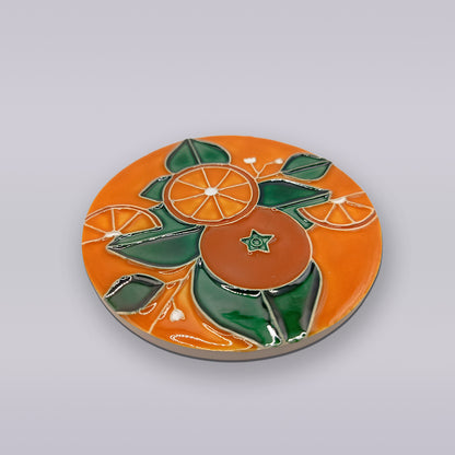 The Algarve Orange Blossom Ceramic Coaster by Miradouro Ceramics features a glossy orange surface with a raised orange fruit, slices, and green leaves, inspired by Algarve coaster designs and shown on a plain light gray background.