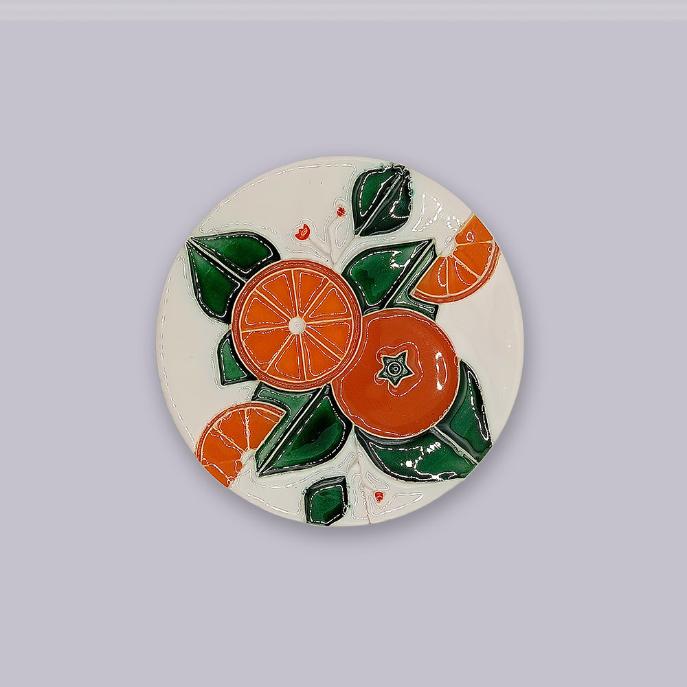 The Algarve Orange Harvest Ceramic Coaster by Miradouro Ceramics features hand-painted orange slices, a whole orange, green leaves, and red dots on white. The coaster is shown resting on a light gray surface.