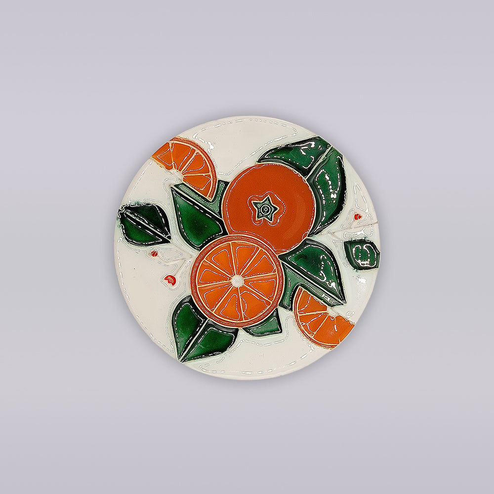 The Algarve Orange Harvest Ceramic Coaster by Miradouro Ceramics features hand-painted orange slices and green leaves on glossy white ceramic, with vibrant artwork centered on a plain light gray surface.