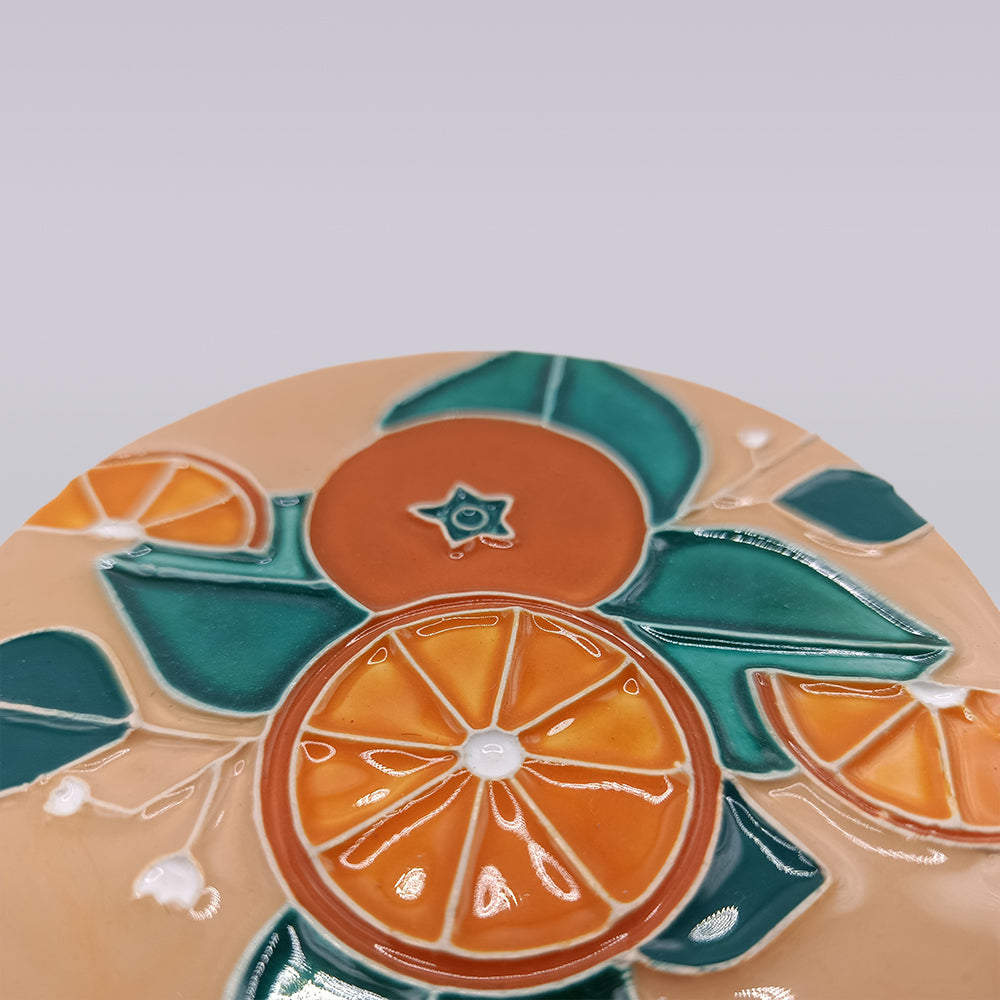 The Algarve Sandstone Orange Ceramic Coaster by Miradouro Ceramics features embossed orange slices, green leaves, and white accents on beige, inspired by Algarve styles. Its colorful, glossy artwork has a smooth finish.