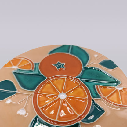 The Algarve Sandstone Orange Ceramic Coaster by Miradouro Ceramics features embossed orange slices, green leaves, and white accents on beige, inspired by Algarve styles. Its colorful, glossy artwork has a smooth finish.