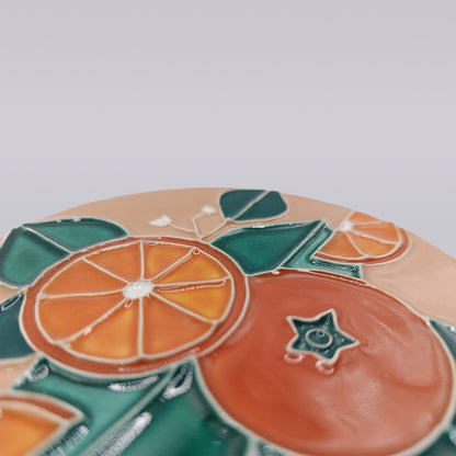 A close-up of the Algarve Sandstone Orange Ceramic Coaster by Miradouro Ceramics, showcasing embossed orange slices and green leaves on a smooth, light surface.