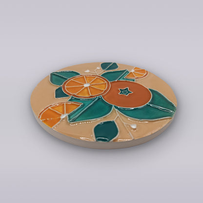 The Algarve Sandstone Orange Ceramic Coaster by Miradouro Ceramics features a glossy, vibrant raised design of orange slices and green leaves on smooth beige ceramic.