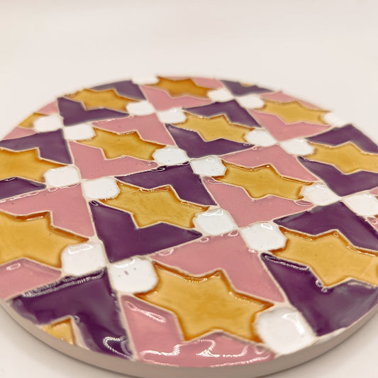 The Azulejo Pop coaster by Miradouro Ceramics is a round handmade ceramic tile with glossy finish, featuring a geometric pattern of yellow stars on alternating pink, purple, and white backgrounds.