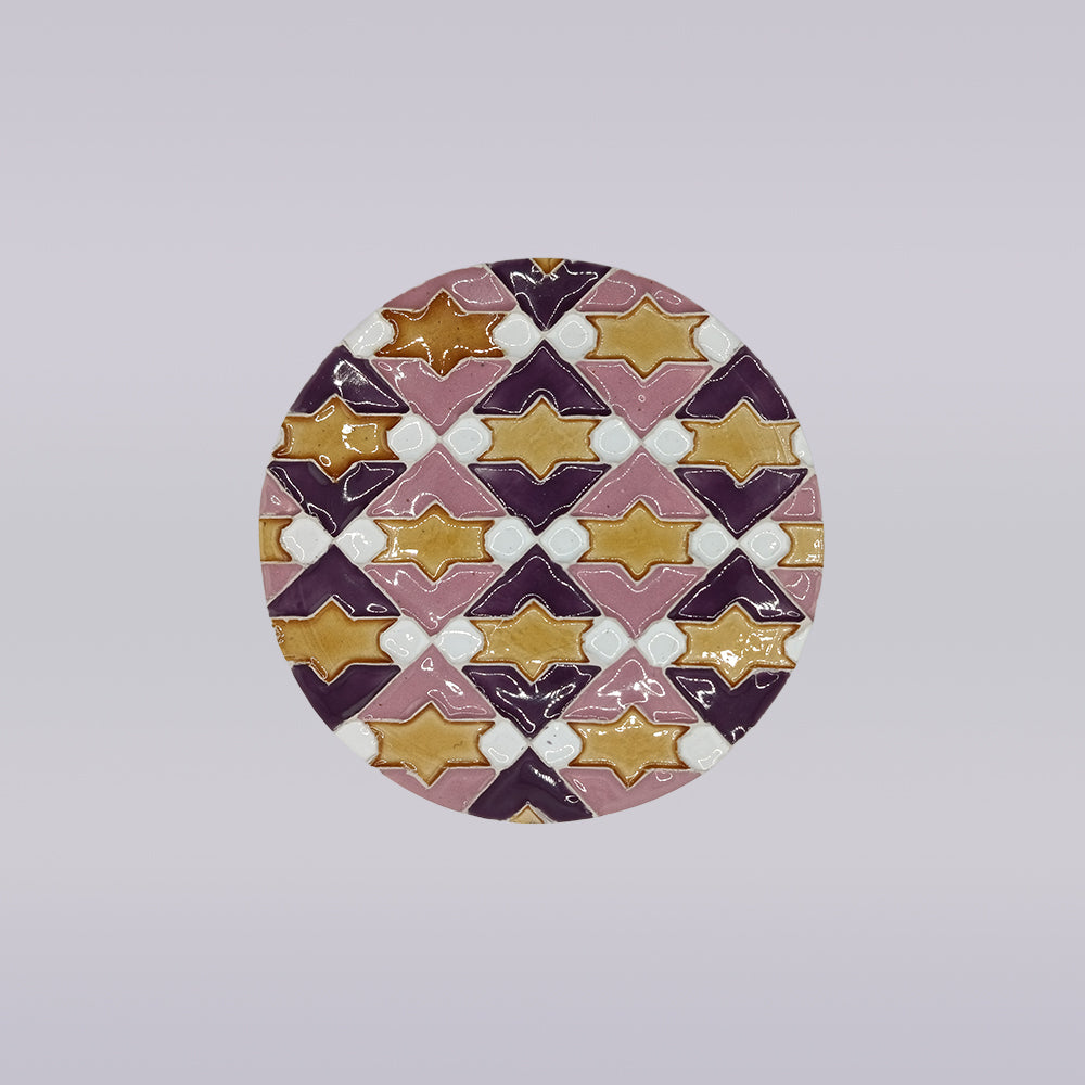 The Azulejo Pop by Miradouro Ceramics is a round handmade ceramic tile coaster featuring a geometric pattern of interlocking diamonds and star shapes in purple, gold, pink, and white, inspired by Portuguese ceramics on a light gray background.