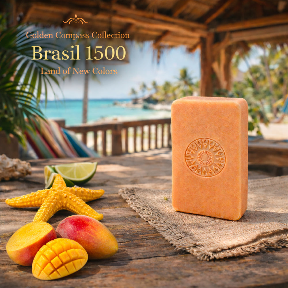 A rectangular orange 100ml Brasil 1500 – Land of New Colors soap bar with a compass design rests on a rustic table next to sliced mango, star fruit, and lime, evoking exotic Brazilian scents beside a tropical beach scene.