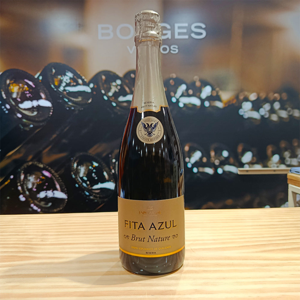 A bottle of Borges Wines Fita Azul Brut Nature Sparkling Wine Reserve rests on a wooden surface, its gold label and foil shining, ideal for Gouveio lovers in a wine shop or cellar with rows of bottles blurred behind.