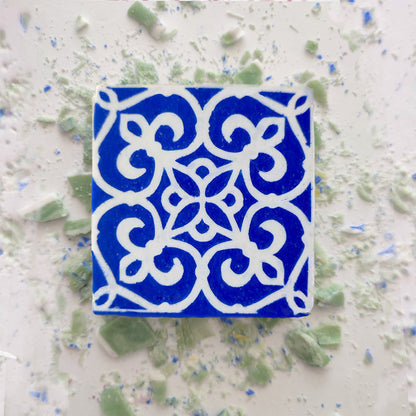 A blue Mosaico Azul Green Tea Soap by Lisbão, featuring a detailed white floral and geometric pattern, rests on a white surface dotted with green and blue fragments, evoking handcrafted Portuguese azulejos.