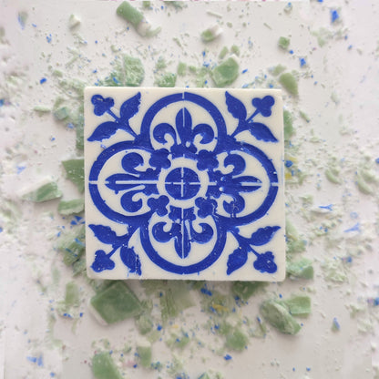 A square Mosaico Azul Green Tea Soap by Lisbão, featuring a blue floral and geometric pattern inspired by Portuguese azulejos, rests on a white surface surrounded by scattered green and blue soap fragments.