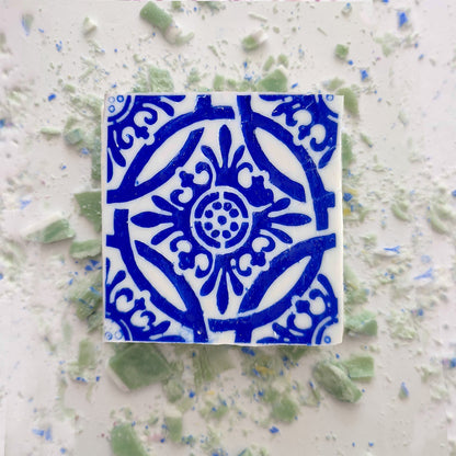 The Mosaico Azul Green Tea Soap by Lisbão features an intricate blue pattern on white, inspired by Portuguese azulejos, and is shown on a white surface with scattered green and blue ceramic fragments.