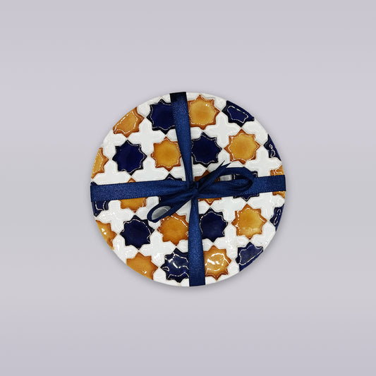 The Heritage Star Mosaic Coaster by Miradouro Ceramics features a Mudéjar star pattern in white, navy blue, and mustard yellow, tied with a dark blue ribbon, displayed on a light gray background.