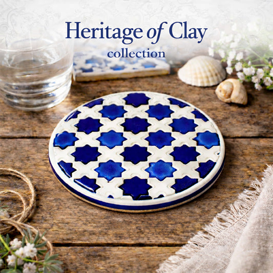 The Heritage Blue Star Mosaic Coaster by Miradouro Ceramics, featuring a blue and white mosaic pattern, rests on a rustic wooden table near a glass of water, shells, flowers, twine, and linen. Text: “Heritage of Clay collection.”.