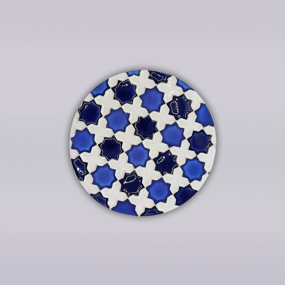 The Heritage Blue Star Mosaic Coaster by Miradouro Ceramics features a round ceramic design with blue and white interlocking stars and crosses, inspired by hand-painted azulejo mosaics, on a light gray background.