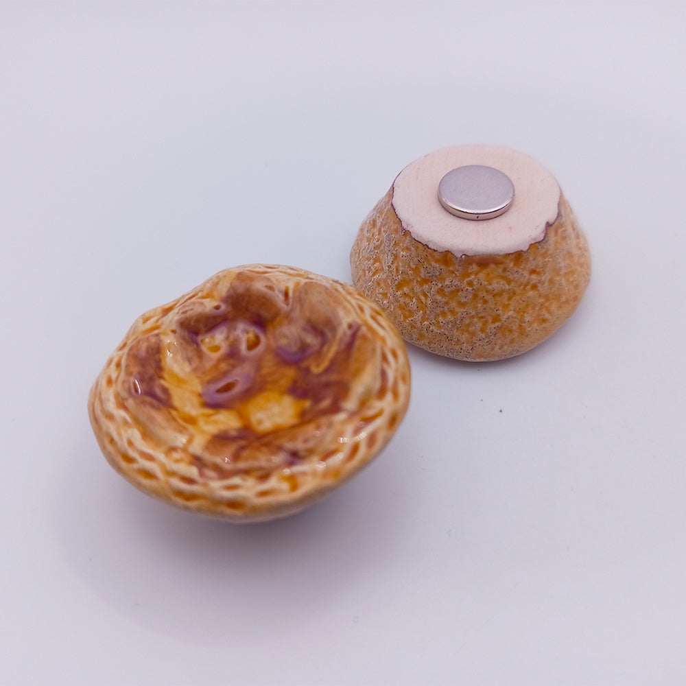 Two Ceramic Belém Custard fridge magnets by Tejo Shop, designed to resemble Portuguese egg tarts, including one positioned upright and the other upside down showing a hidden magnet, are elegantly arranged on a white background. These magnets serve as a charming cultural tribute to Portugal's iconic dessert.