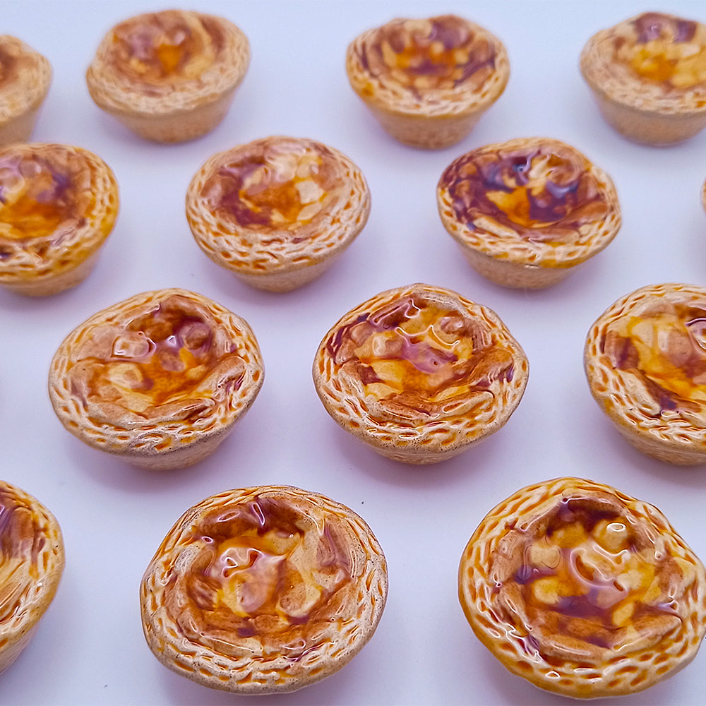 A selection of miniature pastries with glazed, golden-brown tops are arranged in rows on a white background. Similar to the craftsmanship of the Tejo Shop's Ceramic Belém Custard Fridge Magnet, these pastries feature a flaky texture and shiny appearance, embodying cultural artistry.