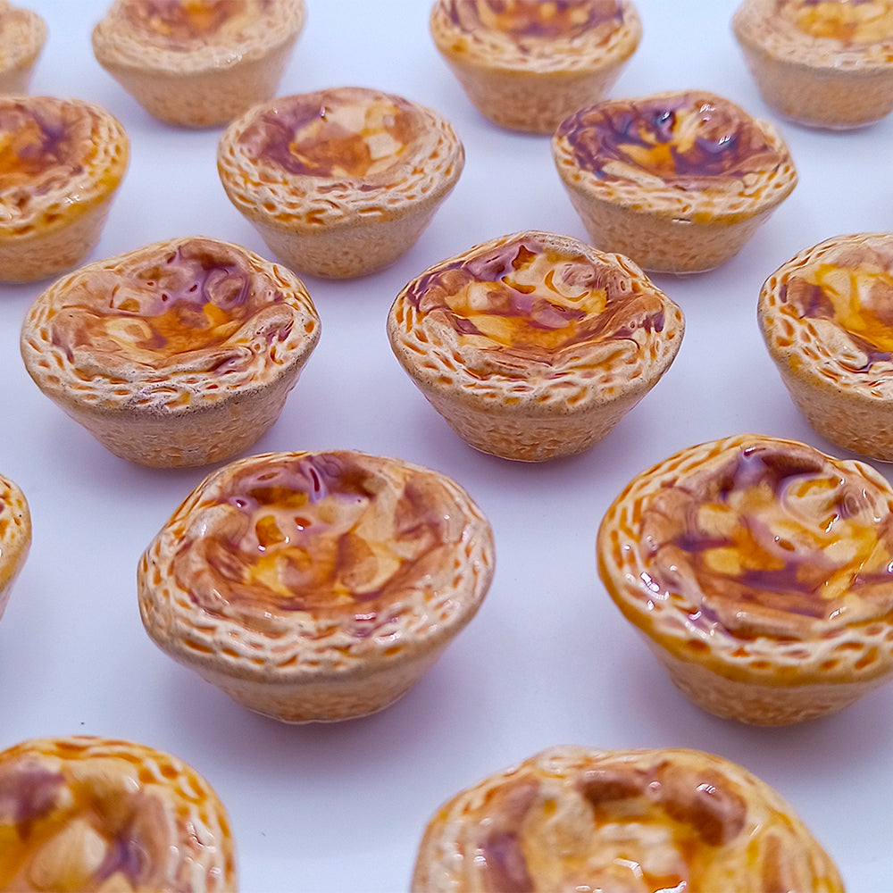 Miniature ceramic pies, showcasing intricate details and a golden-brown crust with a lustrous swirl of filling, are artfully displayed on a white backdrop. These ceramic treasures from Tejo Shop, resembling savory meat pies yet reminiscent of freshly baked goods, capture the essence of Belém custard tarts as delightful cultural icons. Perfectly crafted as fridge magnets under the name "Ceramic Belém Custard," they serve as charming decorative highlights.