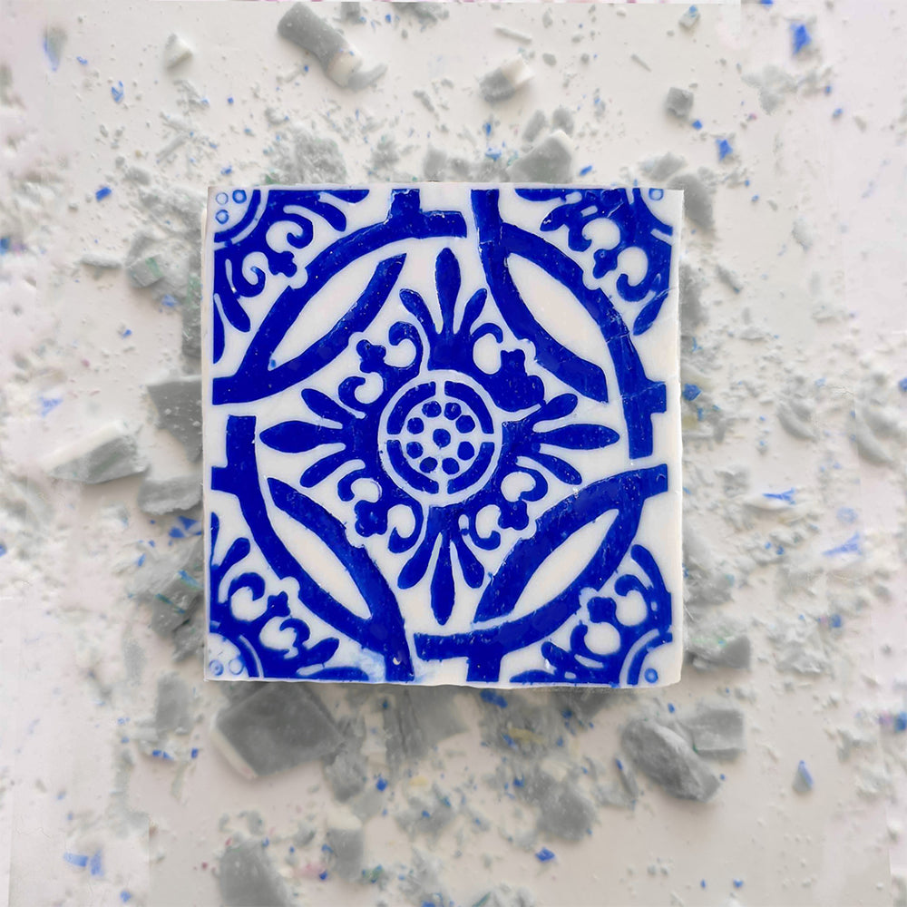 The Jasmine Courtyard Soap by Lisbão sits on a white surface with broken ceramic tiles, echoing the blue floral and geometric artistry of handmade Portuguese tiles.