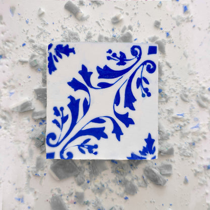 A white ceramic tile with an ornate blue floral pattern rests on a white surface scattered with small, broken pieces—evoking the intricate beauty of Lisbão's Jasmine Courtyard Soap.