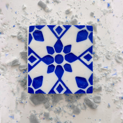 A blue and white geometric floral ceramic tile rests on a white surface with scattered small tiles, capturing the artisanal charm of Lisbão’s Jasmine Courtyard Soap.