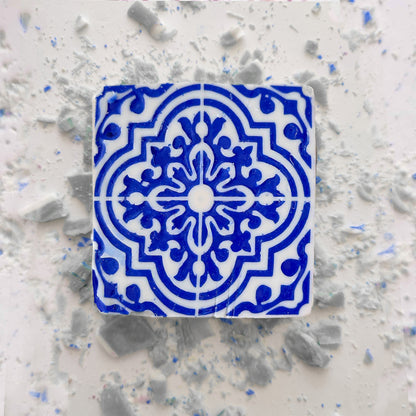 A Lisbão Jasmine Courtyard Soap rests on a light surface beside a decorative blue and white geometric ceramic tile, with scattered dust and broken tile fragments surrounding them.