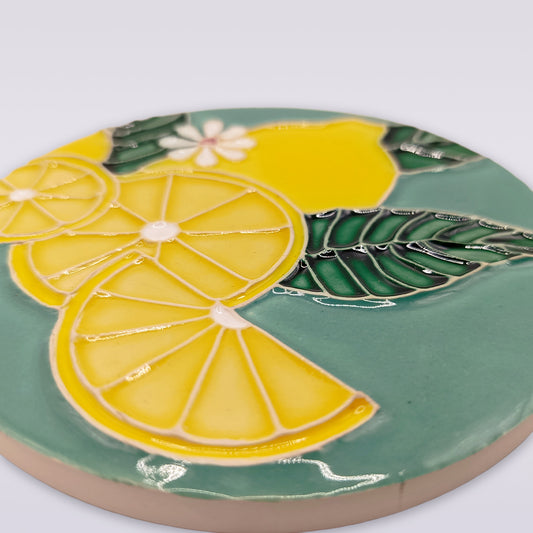 The Lemon Blossom Hand-Painted Ceramic Coaster from Miradouro Ceramics features a raised design with lemon slices, whole lemons, green leaves, and a white flower on a green background, inspired by Portuguese craftsmanship.