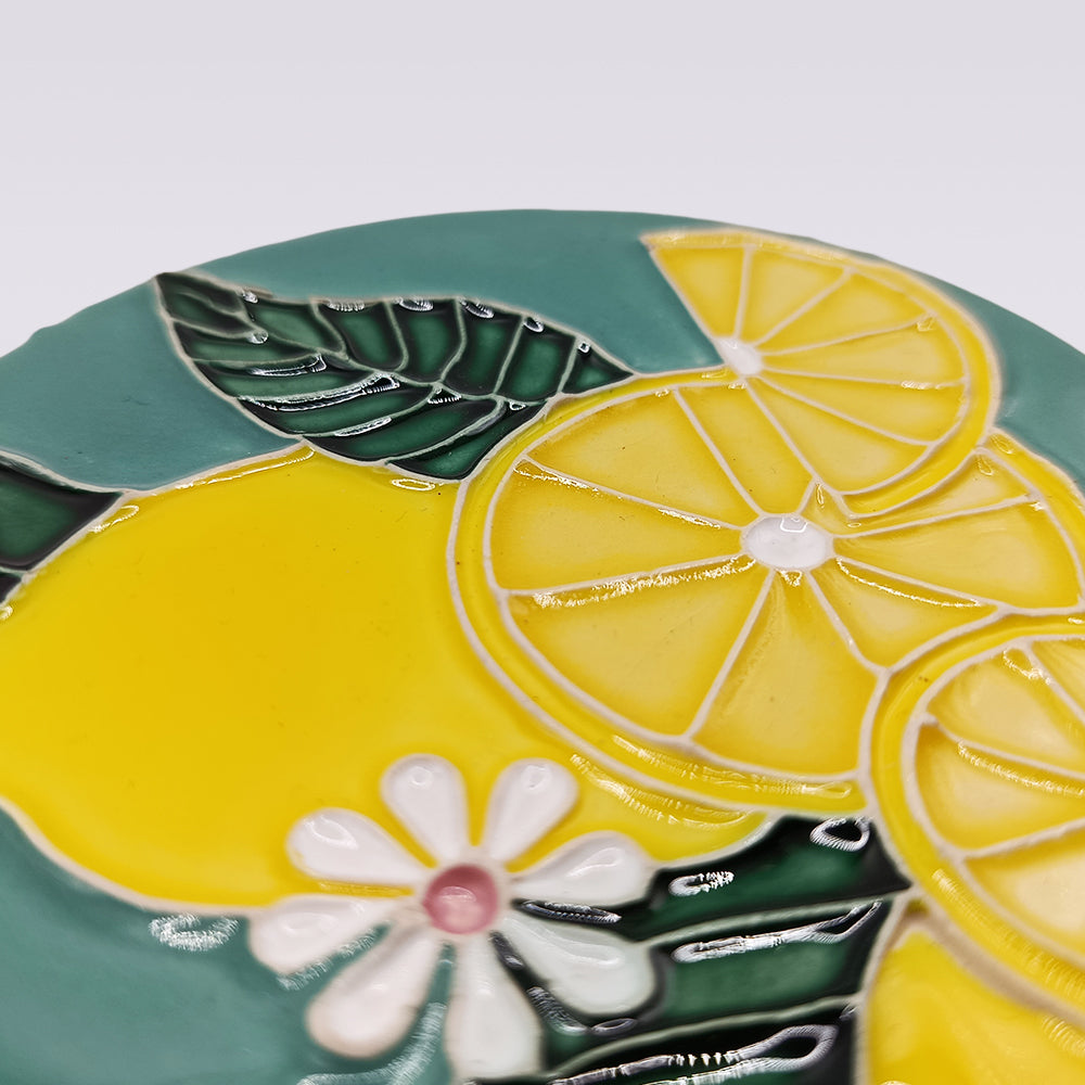 A close-up of Miradouro Ceramics’ Lemon Blossom Hand-Painted Ceramic Coaster, showcasing yellow lemons, green leaves, and white flowers with red centers on a teal background—a must-have for fans of Portuguese artisan homeware.