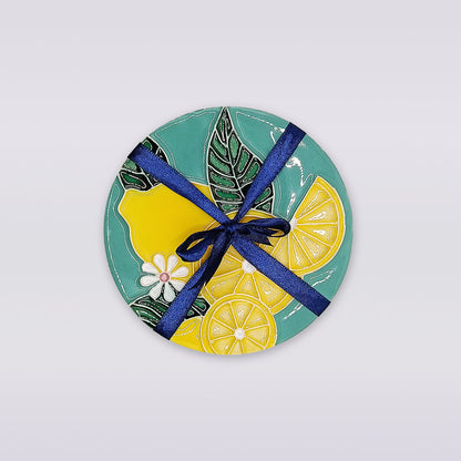 The Lemon Blossom Hand-Painted Ceramic Coaster by Miradouro Ceramics features lemons, white flowers, and green leaves, tied with a dark blue ribbon—a charming piece of Portuguese artisan homeware for your table.