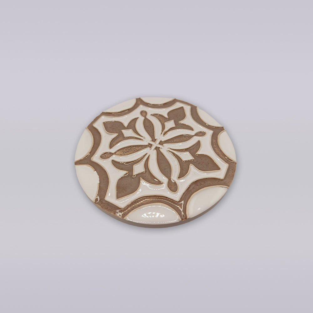 The Lusitana TerraFleur by Miradouro Ceramics is a round handcrafted tile with a raised, symmetrical floral pattern in beige and white, highlighting the elegance of Portuguese ceramics on a light background.