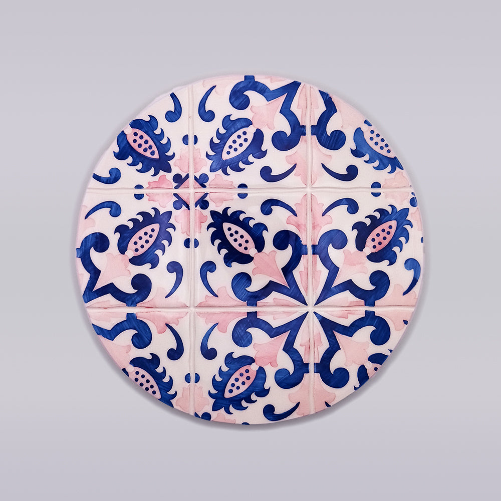 A round plate featuring a vibrant design inspired by traditional Portuguese tiles. The pattern includes blue floral and geometric shapes with pink accents on a white background, creating an intricate symmetrical design. The background is plain and light grey, reminiscent of a Monsaraz Ceramic Kitchen Trivet by Tejo Shop.