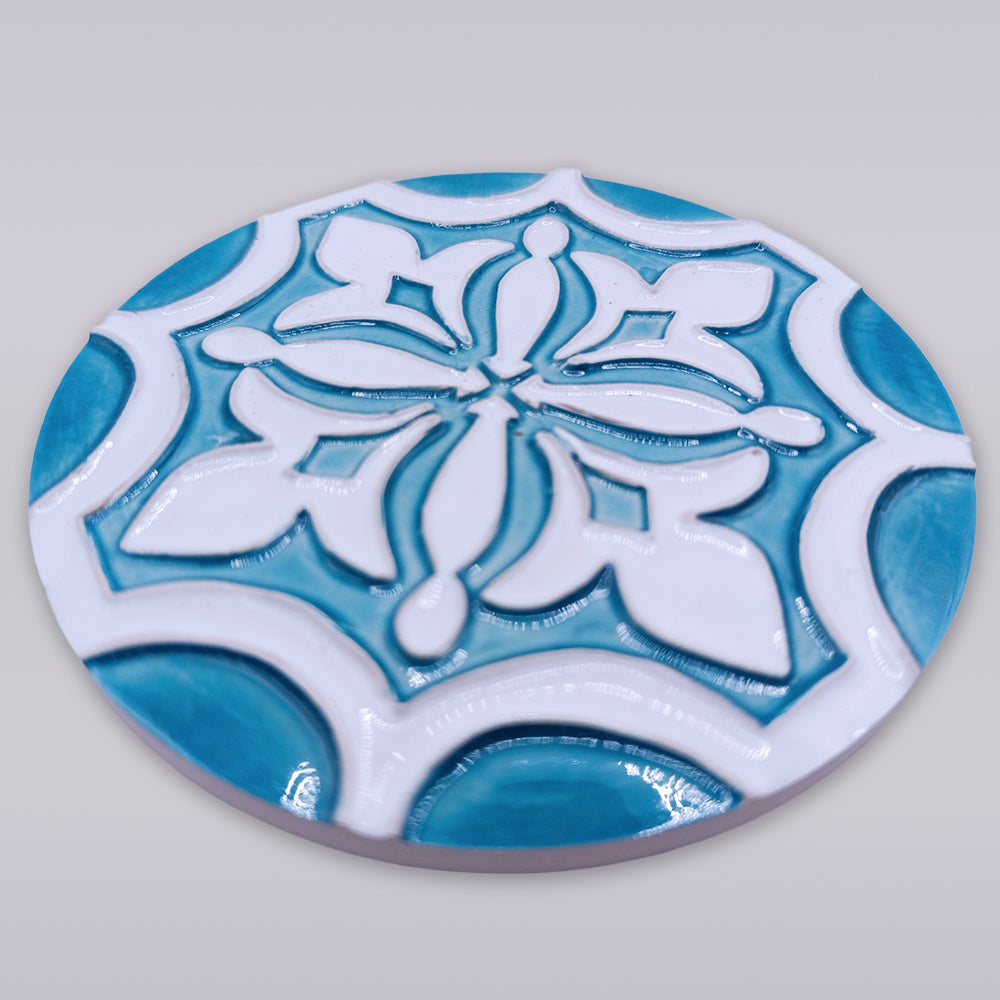 The Rosacrux Atlântica by Miradouro Ceramics is a round, hand-shaped ceramic tile featuring a raised geometric and floral pattern in white and turquoise blue on light gray, with a glossy finish and slightly elevated design.