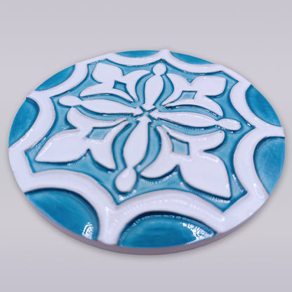 The Rosacrux Atlântica by Miradouro Ceramics is a round, hand-shaped ceramic tile featuring a raised geometric and floral pattern in white and turquoise blue on light gray, with a glossy finish and slightly elevated design.