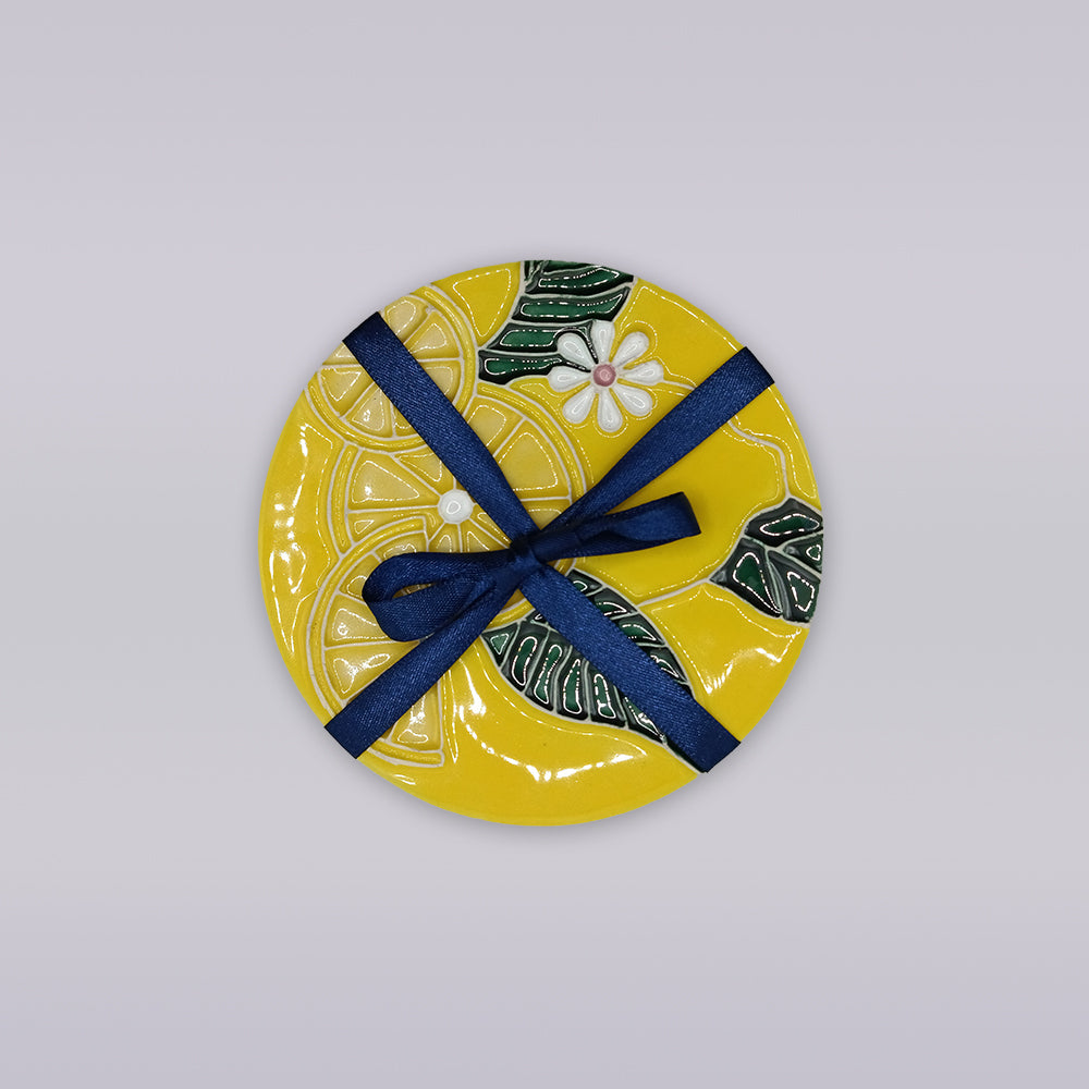 The Tejo Lemon Blossom Coaster – Ribatejo Edition by Miradouro Ceramics is a round yellow coaster with a floral and leaf design, tied with a dark blue ribbon—perfect artisan decor for any space.