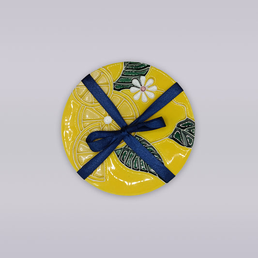 The Tejo Lemon Blossom Coaster – Ribatejo Edition by Miradouro Ceramics is a round yellow coaster with a floral and leaf design, tied with a dark blue ribbon—perfect artisan decor for any space.