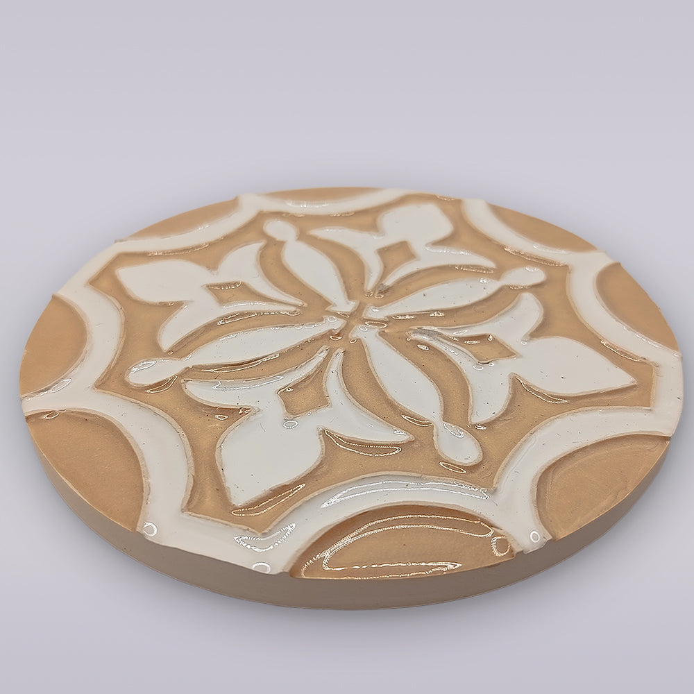 The Terra Manuelina by Miradouro Ceramics is a round, tan ceramic coaster featuring a raised, glossy white floral and geometric design—an artisanal accent for your home decor.