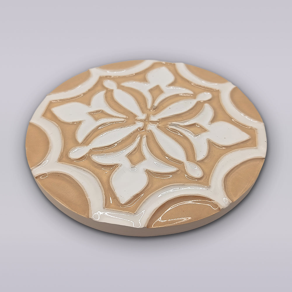 The Terra Manuelina by Miradouro Ceramics is a round, handcrafted ceramic coaster featuring a glossy beige and white geometric floral design inspired by Portuguese ceramics—an artisanal touch for your home decor.