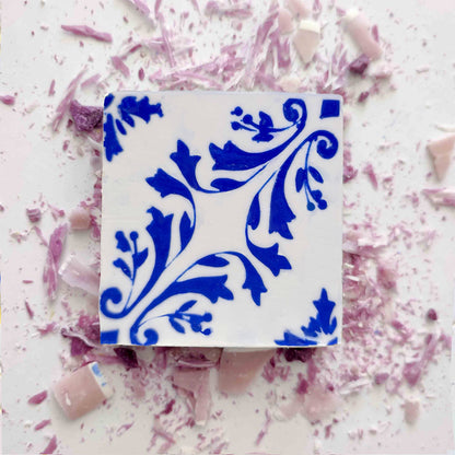 The Lisbão Violet Azulejo Ritual is a square white Portuguese tile soap with ornate blue floral designs, scattered with pink and purple ceramic pieces, evoking a delicate violet scent and artisanal charm.