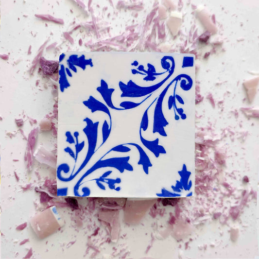 The Lisbão Violet Azulejo Ritual is a square white Portuguese tile soap with ornate blue floral designs, scattered with pink and purple ceramic pieces, evoking a delicate violet scent and artisanal charm.