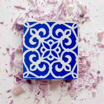 The Lisbão Violet Azulejo Ritual is a square blue tile inspired by Portuguese azulejos, with white floral and geometric patterns, displayed on a surface scattered with pink and purple shards.