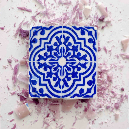 Lisbão's Violet Azulejo Ritual features a square tile with an intricate blue and white pattern, reminiscent of Portuguese design, surrounded by scattered pink and purple shards.