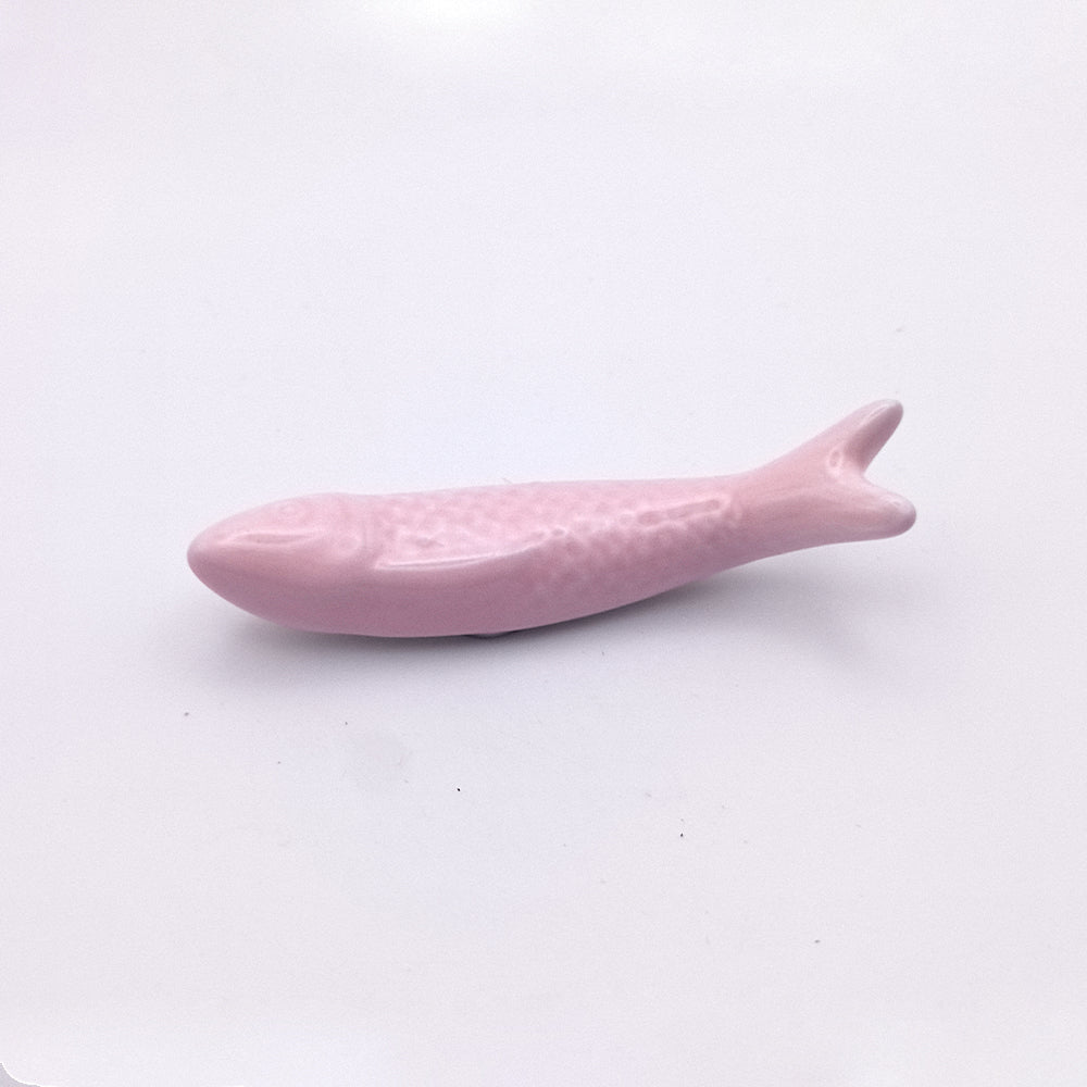 A small, smooth, pink fish figurine lies on a white surface. This Ceramic Sardine Fridge Magnet by Tejo Shop features a simple design with basic details such as fins, scales, and a tail—perfect for adding a touch of kitchen decor.