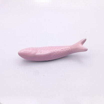 A small, smooth, pink fish figurine lies on a white surface. This Ceramic Sardine Fridge Magnet by Tejo Shop features a simple design with basic details such as fins, scales, and a tail—perfect for adding a touch of kitchen decor.