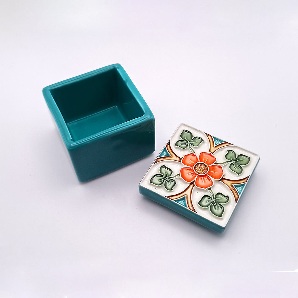 A small square teal Barcelos Ceramic Box from Tejo Shop with a removable lid featuring a colorful floral design. The lid has a central orange flower surrounded by green leaves and blue triangular accents. This hand-painted ceramic piece sits elegantly on a plain white surface, adding sophisticated home decor to any space.