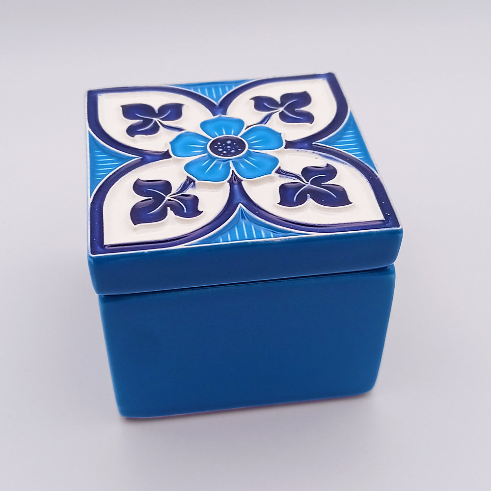 A small, square ceramic box with a vibrant blue base. The lid features a hand-painted flower motif with four petals and surrounding white and dark blue designs, creating a symmetrical, decorative pattern. This lavish Batalha Ceramic Box by Tejo Shop is both elegant and eye-catching.